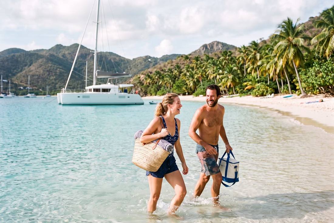 Couple wading ashore from a chartered catamaran anchored off a palm-backed Caribbean beach